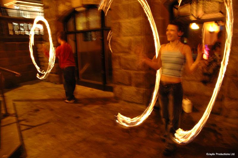 Two people spin fire poi at night under a stone archway, leaving long trails of light