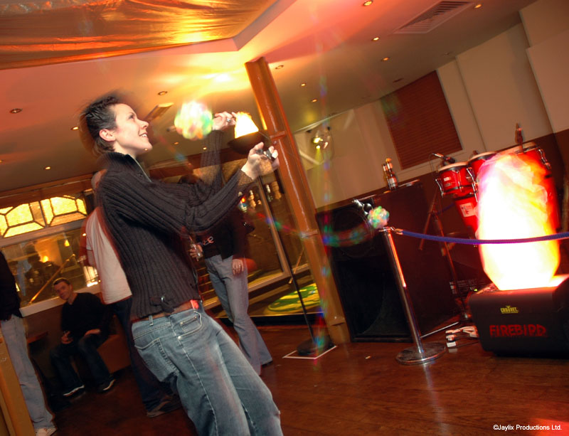 A person in a dark striped top plays with glowing poi beside a band area and a flame effect unit