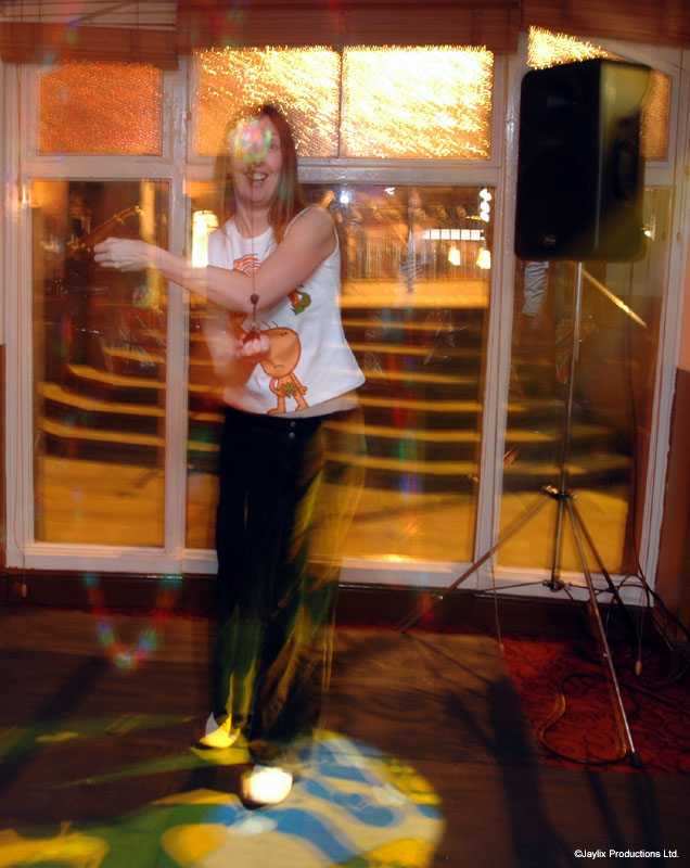 A person in a white top spins glowing poi in front of tall windows and a speaker stand