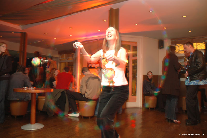 A person in a white top dances with glowing poi in a bar while others sit and stand behind