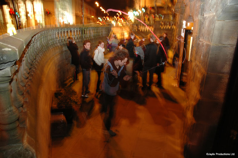 Blurred people walk along a curved stone balustrade at night, with light trails in the distance
