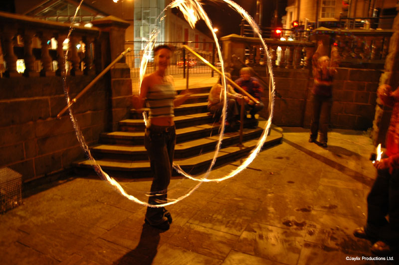 A woman swings two sticks to trace a large ring of light on a stone terrace at night, people seated on steps behind