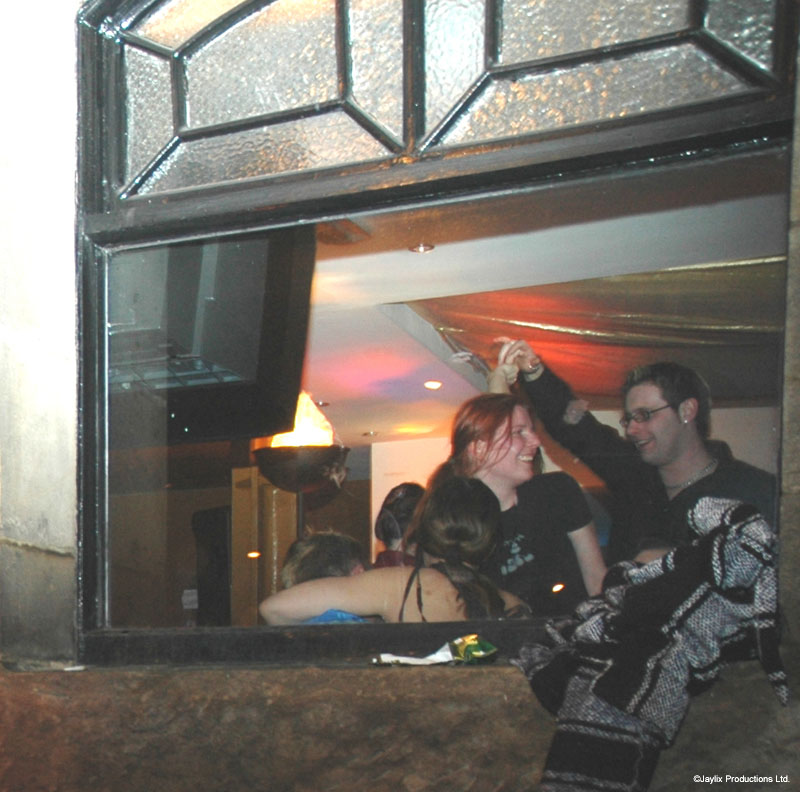 Two people sit together in a window alcove seen from outside, with a flame torch inside the bar