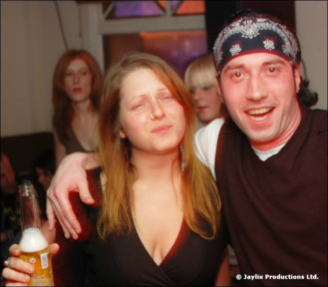 A woman with her eyes half closed and a man in a patterned bandana pose together, the man holding a bottle