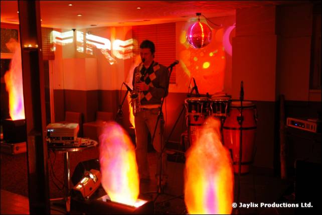A saxophonist plays on a small stage beside congas and two tall flame effects, with a FIRE sign behind
