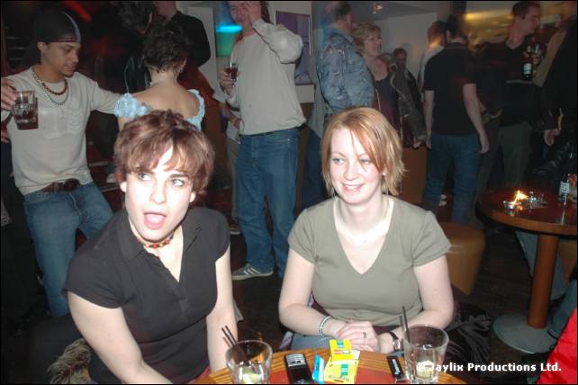 A man and a woman sit at a table with drinks in a busy bar, the man with his mouth open