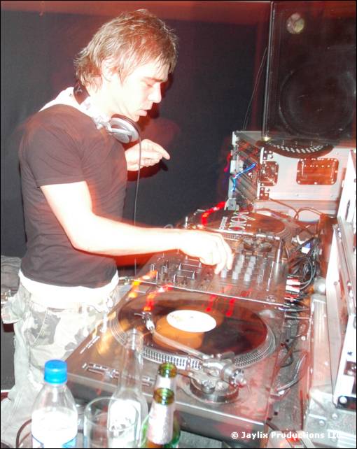 A DJ with spiky blonde hair and headphones mixes at turntables and a mixer, with bottles in the foreground