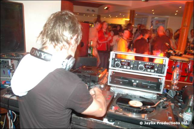 A DJ in a hooded top and headphones plays records at turntables, with a crowd behind