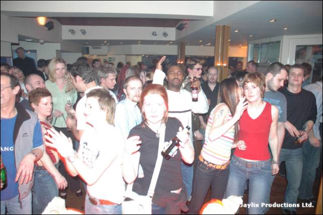 A packed crowd stands and dances in a bar, with a woman in a red top at the centre