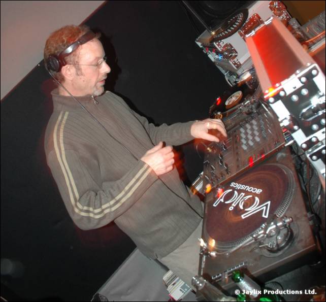 A DJ with glasses and headphones in a striped tracksuit top mixes at a Void Acoustics turntable