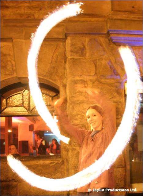 A woman stands inside a large circle of fire trails in front of a stone building with a lit window