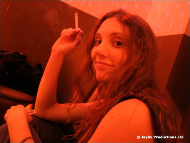 A woman with long hair holds a cigarette and smiles at the camera in red light