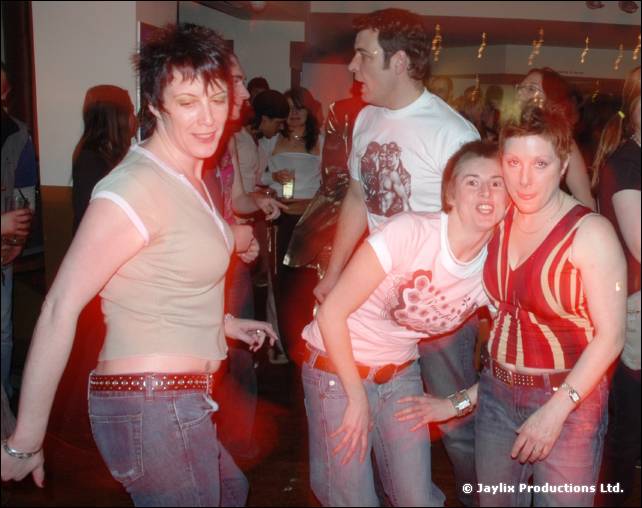 Four people dance and pose together in a bar, two women in front with their arms around each other