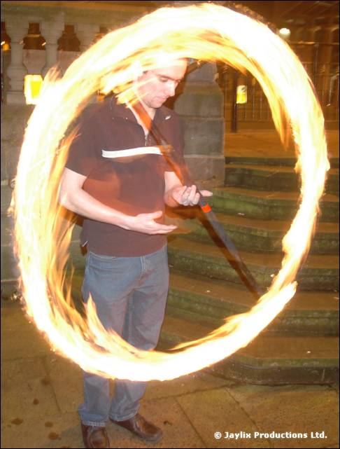 A person in a dark T-shirt stands on steps spinning a staff that leaves a large loop of fire