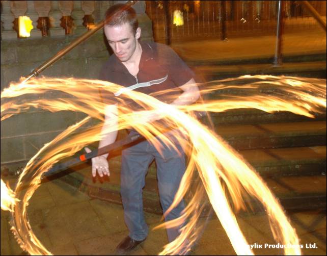 A person in a dark shirt stands on steps with fire trails sweeping around them from a spinning staff