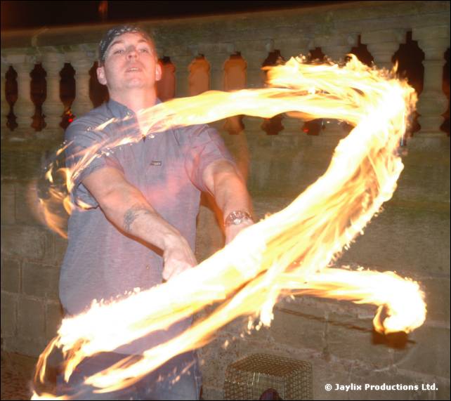 A man in a grey T-shirt swings a long streak of flame in front of a stone balustrade