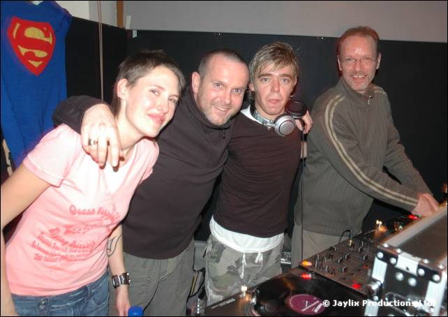 Four people pose behind DJ decks, with a Superman shirt hanging on the wall behind them