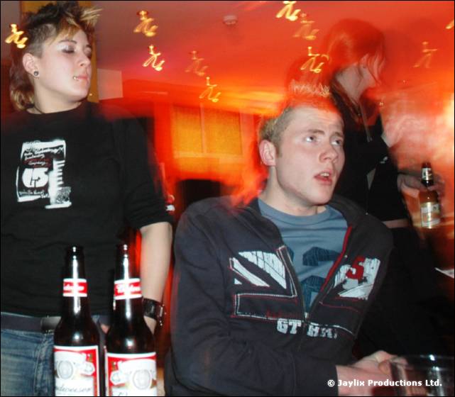 A young man in a dark jacket looks aside beside two beer bottles, with a woman in a black T-shirt behind