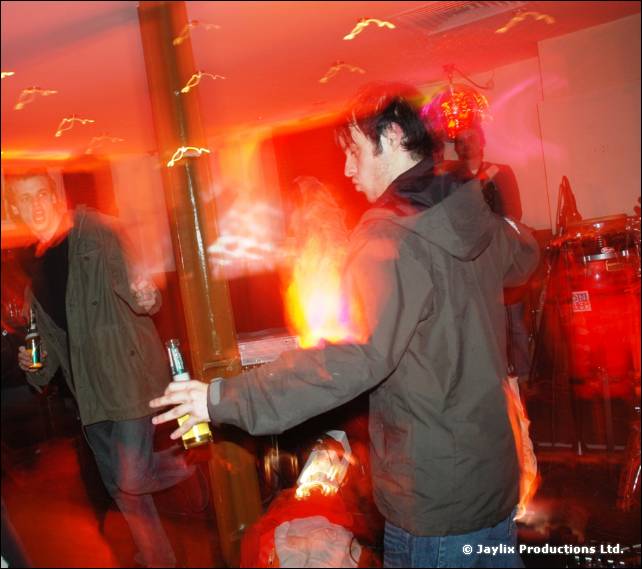 A man in a green jacket holds a bottle in a red-lit bar, with flames and other people blurred behind