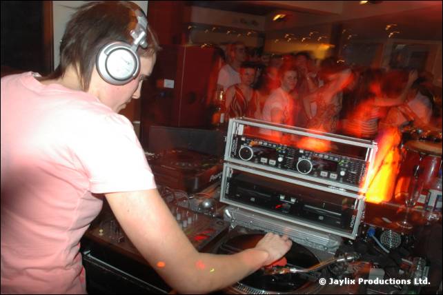 A DJ in a pink T-shirt and headphones mixes at twin CD decks, with a crowd behind