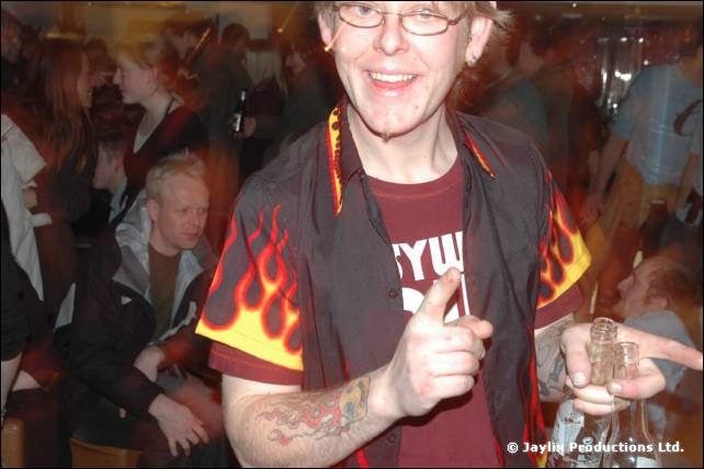 A smiling man with glasses in a flame-patterned shirt points at the camera, with people behind him