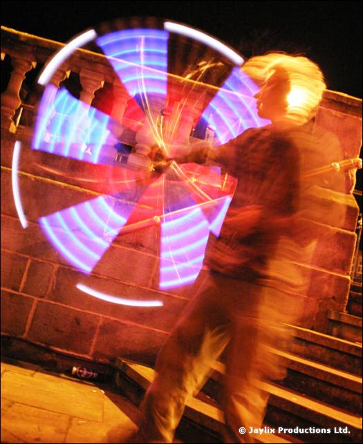 A blurred person spins glowing blue and white poi in front of a stone balustrade at night