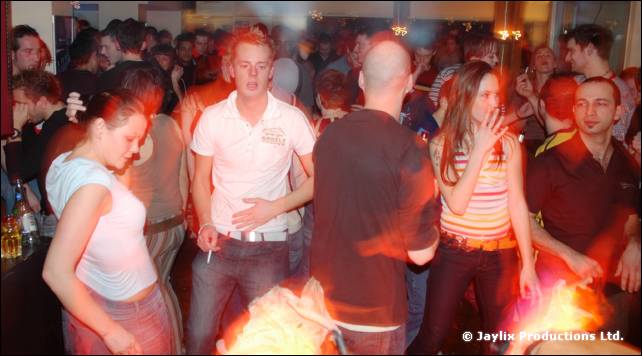 People dance in a crowded bar, including a man in a white shirt with a cigarette and a woman in a striped top