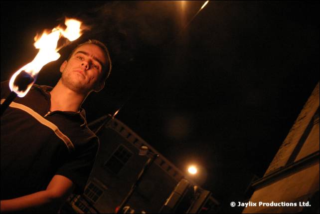 A man in a black polo shirt holds a burning torch at night, a stone building behind him