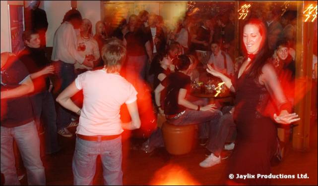 Blurred people dance in a busy bar, with a woman in a dark dress on the right