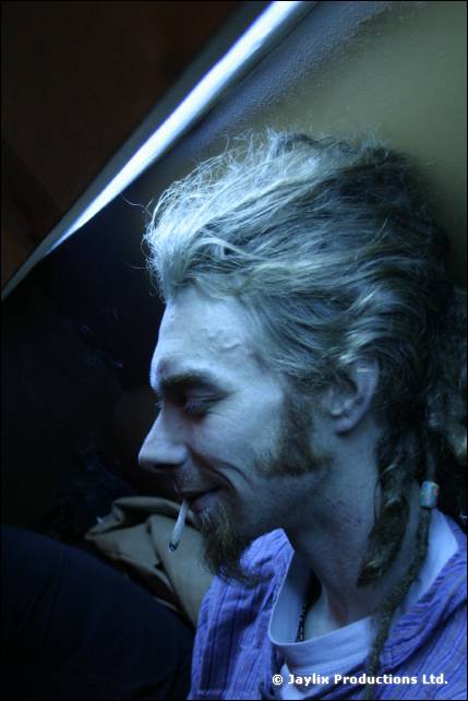 A dimly lit man with greying dreadlocks and a beard looks down, smiling, with a cigarette