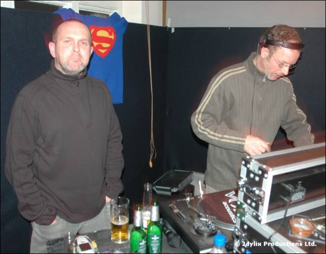 Two men stand by DJ decks, one in a dark jumper and the other in a striped jacket mixing, with a Superman shirt behind