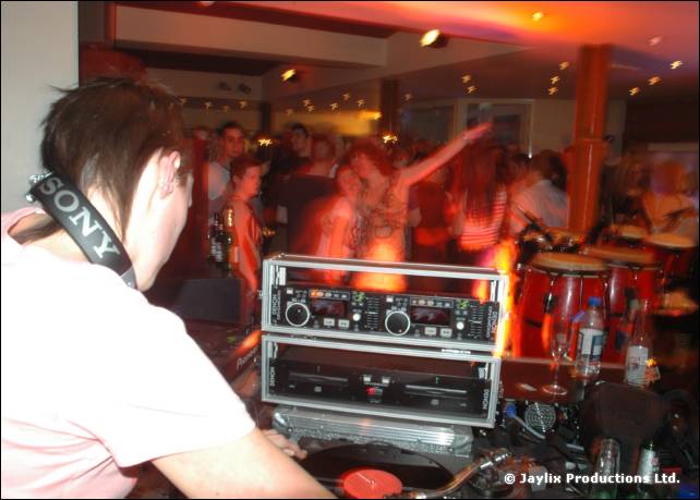 A person in a pink T-shirt wearing a Sony strap stands by twin CD decks, with people dancing behind