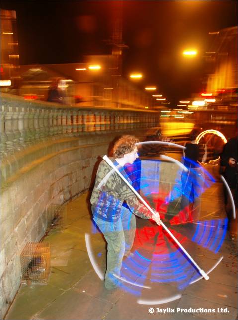 A person spins a white staff with blue and red light trails in a street at night