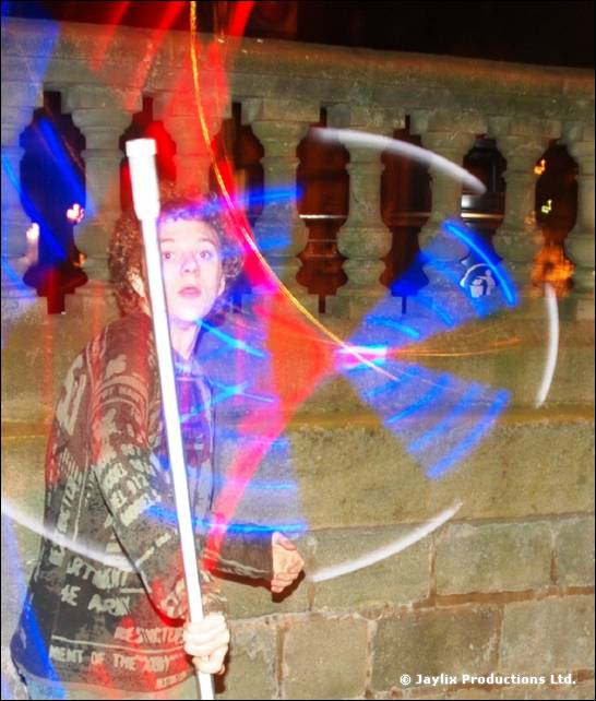 A person with curly hair holds a white staff with blue and red light trails against a stone balustrade