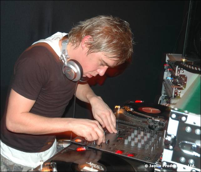 A DJ in a brown T-shirt and headphones mixes at turntables