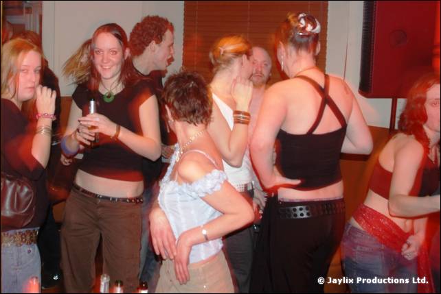 A group of people dance in a bar, including a woman with a drink in a dark top