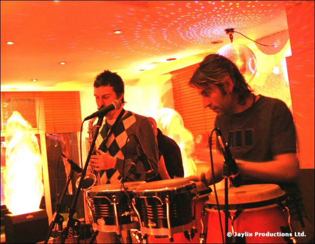 Two musicians play a saxophone and congas on a small stage with a glowing flame effect and mirror ball above