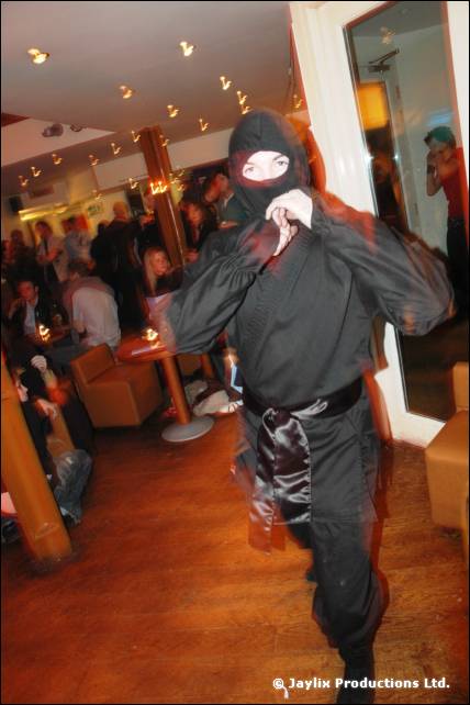 A person dressed as a ninja in black stands in a bar with hands raised