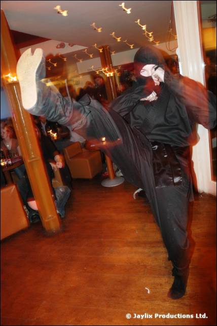 A person dressed as a ninja in black performs a high kick in a bar