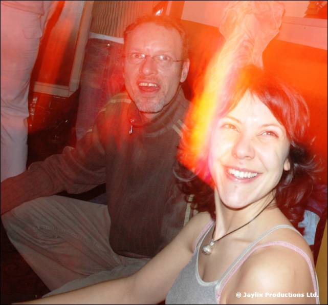 A smiling woman with red hair and a man with glasses sit close together, with a flame glowing behind her
