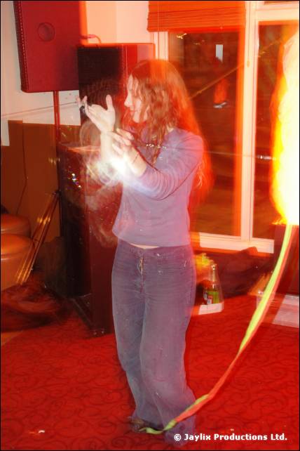 A woman in a blue top and jeans stands with her hands together near a speaker, with a flame at the edge