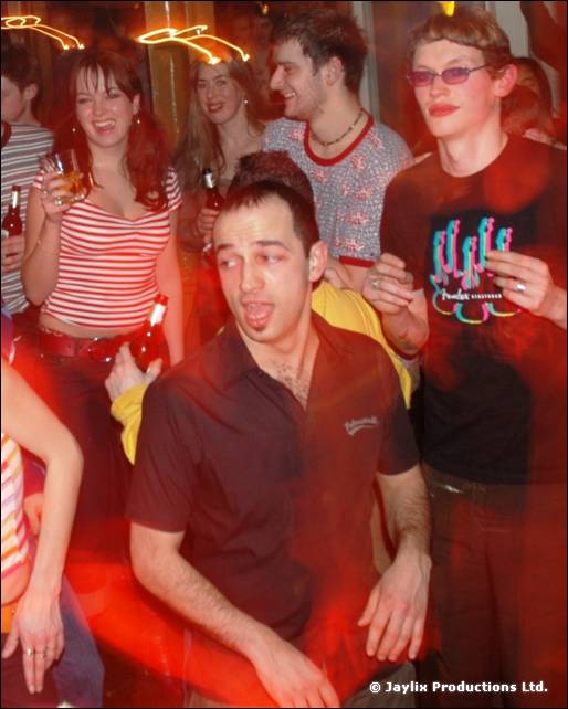 A man in a dark shirt with his mouth open dances among a group in a bar, with a woman in a striped top behind