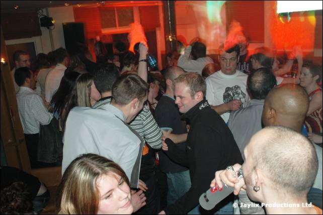 A crowded bar with a woman in the foreground, a bald man holding a microphone, and people dancing behind