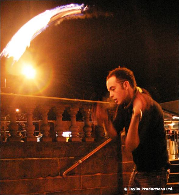 A man in a dark T-shirt swings a flaming prop beside a stone balustrade at night