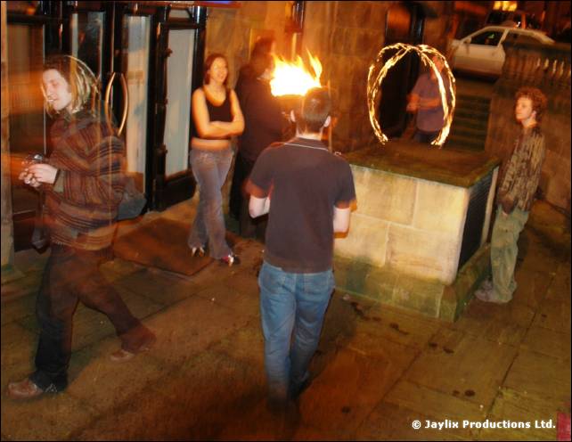 People stand on a street at night by a stone plinth, one holding a flame, with a fire hoop in the background