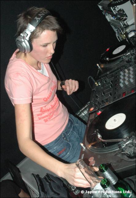 A DJ in a pink T-shirt and headphones leans over turntables, with a bottle on the deck