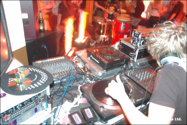 A DJ in headphones plays records at a cluttered set of decks, with flame effects and congas behind