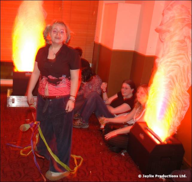 A person stands on a red carpet beside a flame effect, with ribbons at their feet and others seated behind