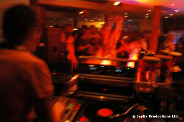 A blurred person in the foreground with decks, congas and dancers lit orange behind