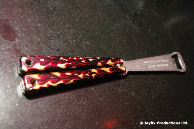 A butterfly knife bottle opener with flame patterned handles lies on a dark surface